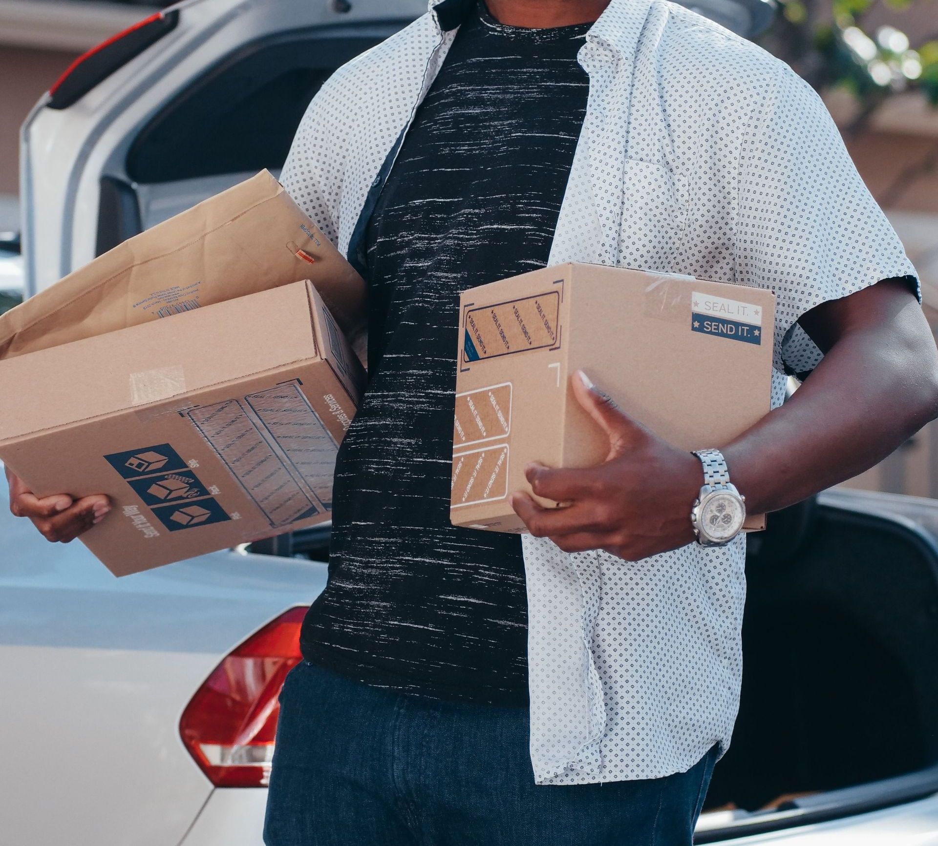 A cheerful man holding packages stands by an open car trunk outdoors, ideal for delivery and logistics concepts.