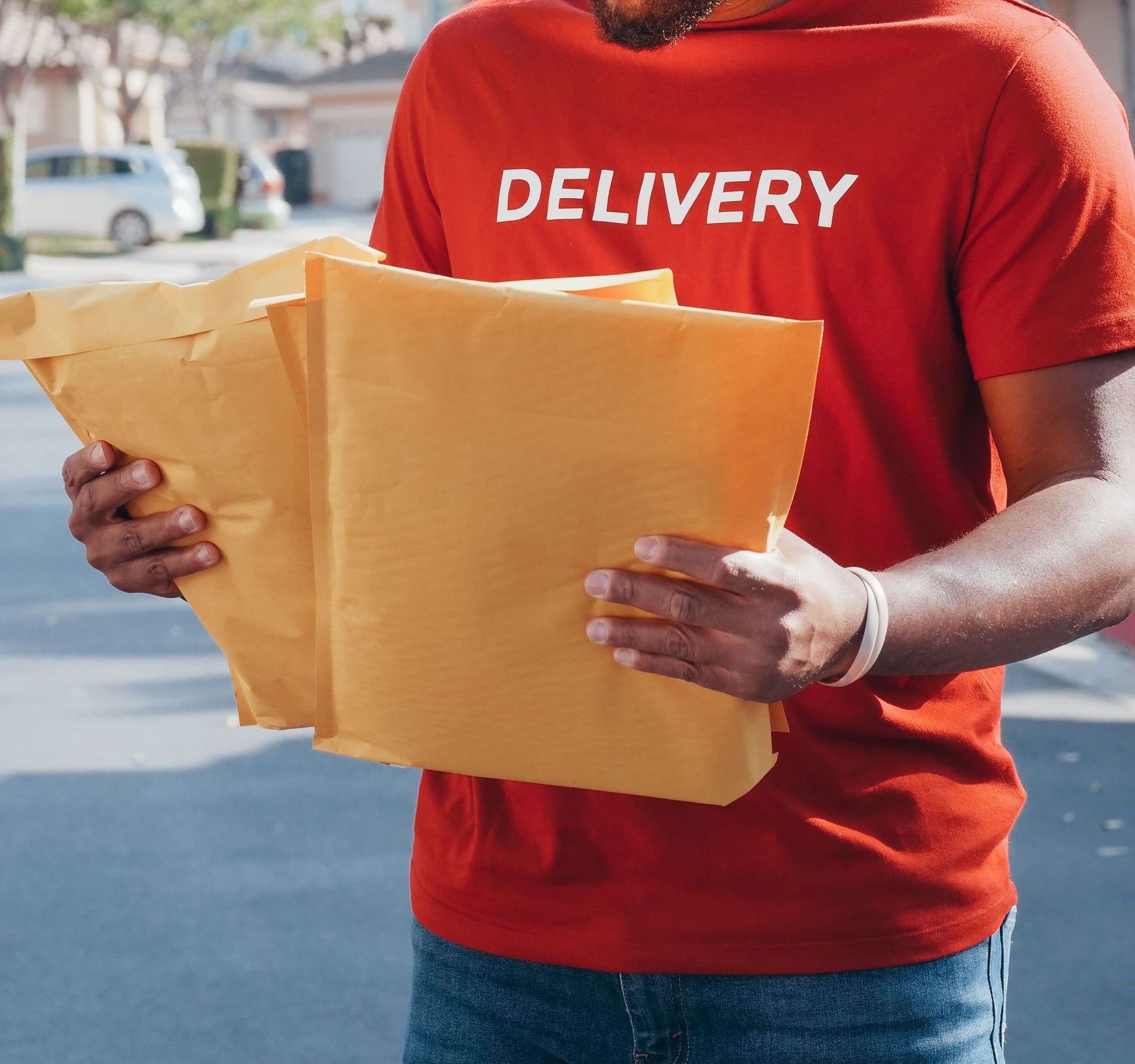 A delivery man in a red uniform holds envelopes while standing outdoors.